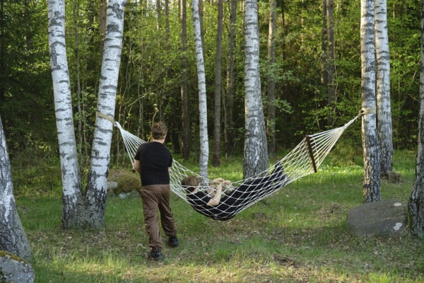Two children enjoy their time outdoors, one relaxing in a hammock suspended between two trees while the other playfully holds onto the hammock