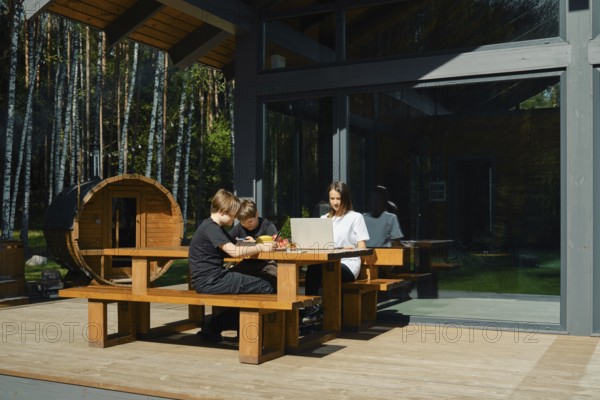 Two children focus on their tasks at a wooden table outside a modern cabin, while their mother guides them using a laptop. Surrounding trees offer a tranquil ambiance perfect for learning