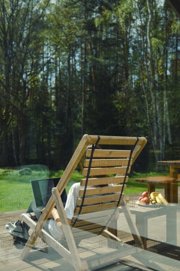 A person enjoys leisure time on a wooden lounge chair, surrounded by picturesque trees and a calm atmosphere. Refreshing fruits lie on a nearby table, enhancing the peaceful outdoor experience