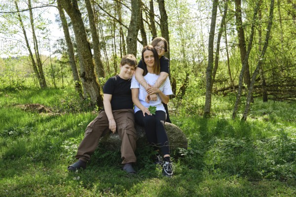 A single mother sits comfortably on a large rock with her two sons in a vibrant green forest clearing. They stopped to rest after a walk in the woods