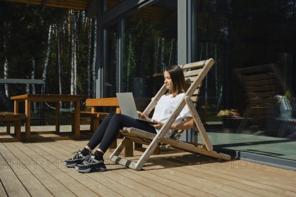 A woman sits comfortably in a wooden chair on a deck, focused on her laptop. Bright sunlight filters through trees, creating a serene atmosphere perfect for remote work or relaxation