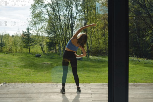 Rear view at a woman tilting her body to the sides on a wooden deck, surrounded by lush greenery and trees.She is stretching her muscles and warming-up before her workout experience