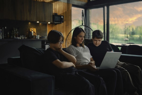 Young mother and her sons watch a series on a laptop sitting in the evening in a cozy living room of a wooden cabin