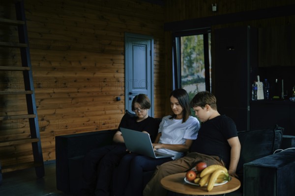 Three children sit closely together on a sofa in a wooden cabin, focused on a laptop. Natural light fills the room, creating a warm atmosphere as they engage in their activity