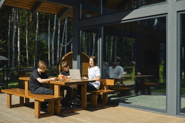 Two children and their mother are seated at a wooden table outside a modern building. They are engaged with digital gadgets and snacks, surrounded by trees, enjoying a calm day in nature