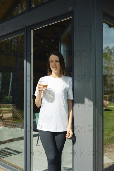 A young woman stands leisurely at the entrance of a stylish house, holding a drink in her hand. Sunlight shines through the glass doors, creating a warm atmosphere in a green backyard