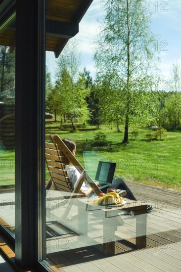 A person enjoys a peaceful moment outdoors, seated in a wooden lounge chair. Surrounded by lush greenery, she is focused on laptop, absorbing the serene atmosphere of nature