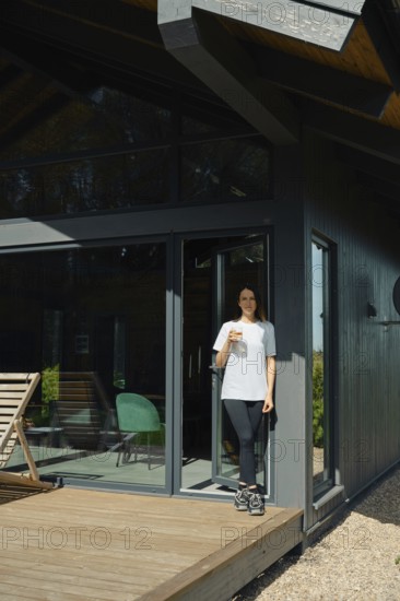 A woman stands on the wooden deck of a stylish cabin, holding a drink while enjoying the serene atmosphere