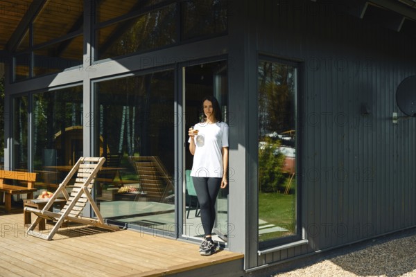 A woman stands on the deck of a stylish house, holding a drink while enjoying a sunny day. The home features large windows and a relaxing outdoor area with furniture, surrounded by greenery
