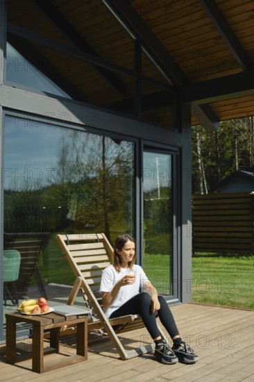 A woman sits comfortably in a wooden lounge chair on a spacious deck, enjoying a refreshing drink