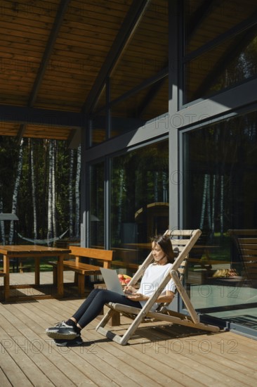A woman sits on a wooden chair outdoors and typing on a laptop. She enjoys her vacation in modern wooden cabin amidst the trees
