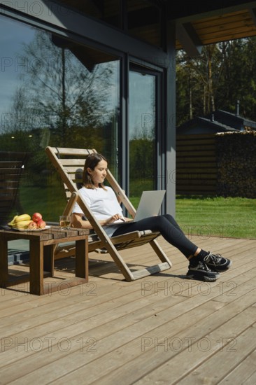 A woman sits comfortably on a deck chair, focused on her laptop. She is scrolling through social media and share moments of her vacation with friends
