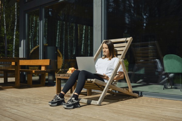 A woman is sitting in a wooden lounge chair, engaged with her laptop on a sunny deck. She enjoys a relaxed atmosphere surrounded by greenery, with fresh fruit nearby and large windows in view