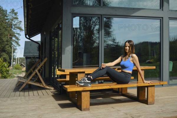 A woman is seated on a wooden bench, stretching her leg while practicing yoga in a serene outdoor setting