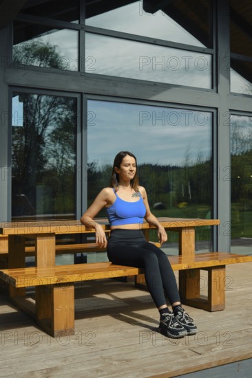 A woman sits on a wooden bench in front of a large glass cabin, basking in natural light. Surrounded by greenery, she wears a blue top and black leggings, appearing relaxed and peaceful
