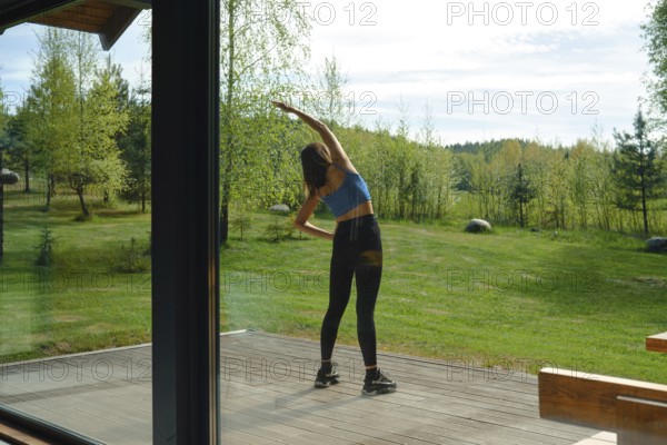 The view from inside the house at a woman is stretching her body on a wooden deck surrounded by lush green trees on a sunny day. The natural setting provides a peaceful backdrop for her fitness activity