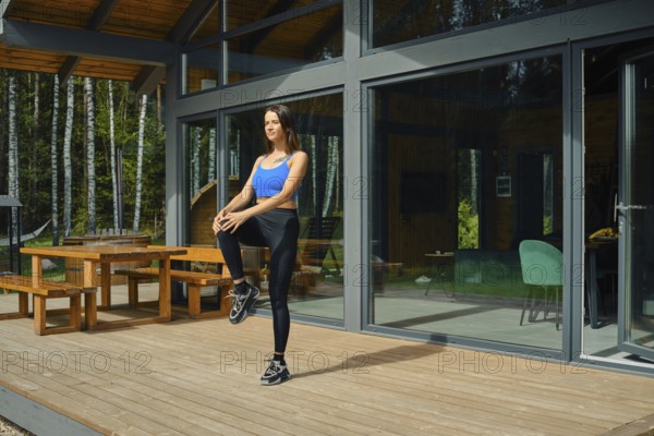 A woman wearing athletic attire is engaged in a warm-up exercise, raising one knee while standing on a wooden deck outside a contemporary cabin. Lush green trees provide a peaceful backdrop