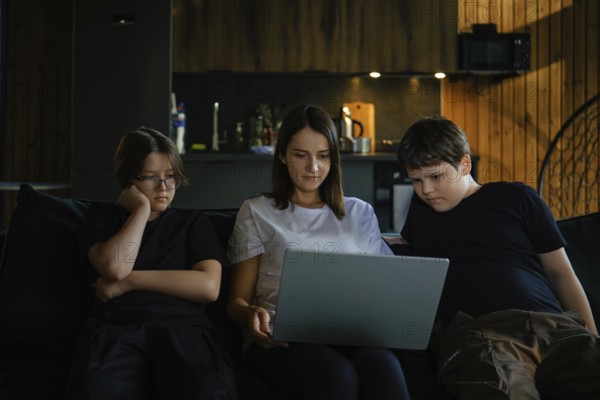 A woman and two children, are sitting closely on a sofa. They focus on a laptop screen in a warmly lit living room. The ambience suggests a relaxed evening activity together
