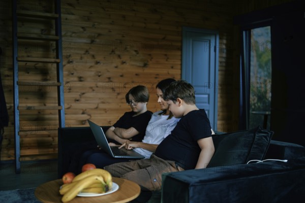 A family sits together on a comfortable couch in a wooden living room, focusing on a laptop. The mother explains the rules and teaches the children using interactive learning