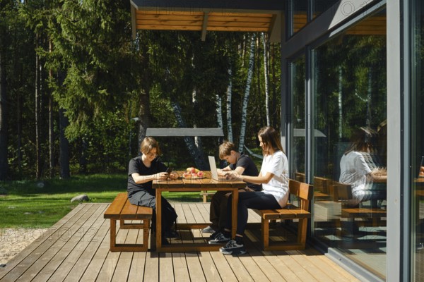 A single mother works remotely at a laptop sitting at a table on the terrace of a country house, and her sons are passionate about a game on a smartphone