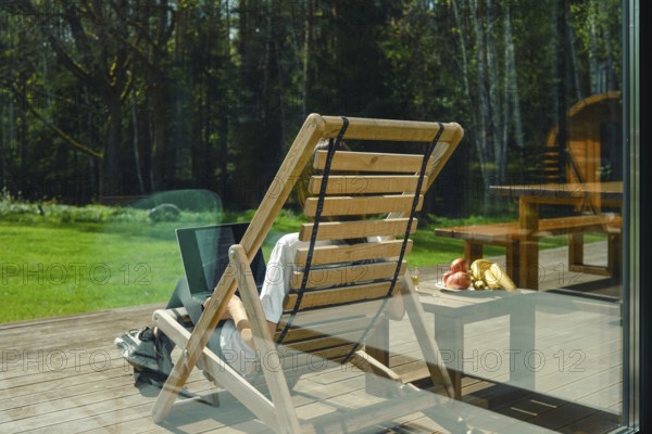 A person works on a laptop while reclining in a wooden lounge chair on a deck surrounded by trees. Fresh fruits are placed nearby, providing a healthy boost in a tranquil environment
