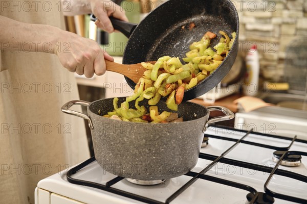 A person transfers sliced vegetables in a pot while adding more ingredients from a frying pan on a gas stove. The kitchen features warm colors and a homely atmosphere with kitchenware in the background