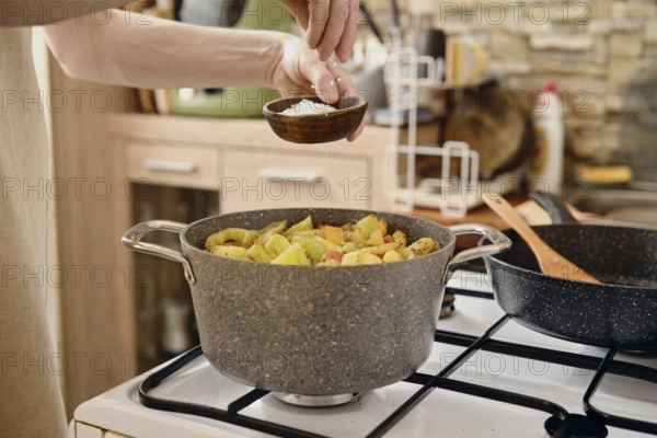 A person is preparing a healthy vegetable dish, adding salt from a small bowl into a pot on a stove. The kitchen features wooden cabinets and cooking utensils scattered on the counter