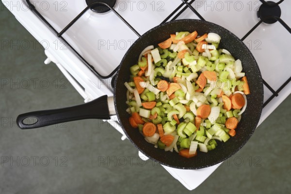 A skillet filled with freshly chopped onions, carrots, and celery sits on a gas stove. The vibrant colors of the vegetables highlight the preparation of a healthy meal in a home kitchen