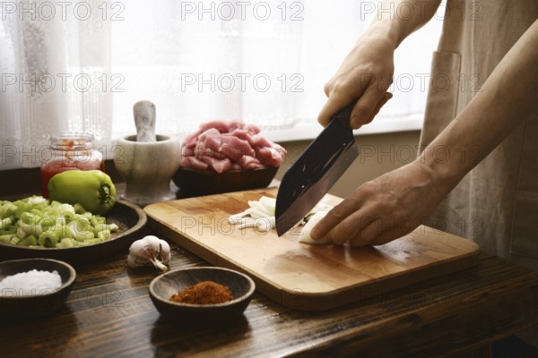 A chef finely chops onion on a wooden cutting board in a warm kitchen. Fresh ingredients like celery and peppers are ready for a flavorful ragout