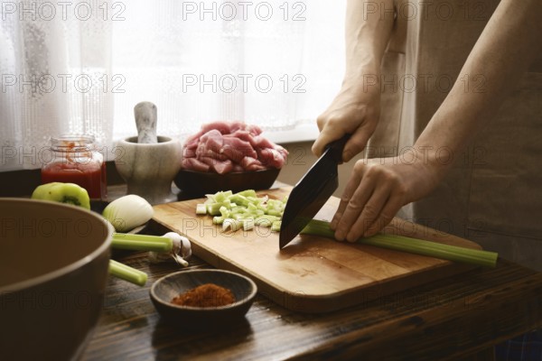 A cook prepares ingredients by chopping celery on a wooden cutting board. Fresh vegetables and meats are arranged on a table, creating a warm, inviting kitchen atmosphere for meal preparation