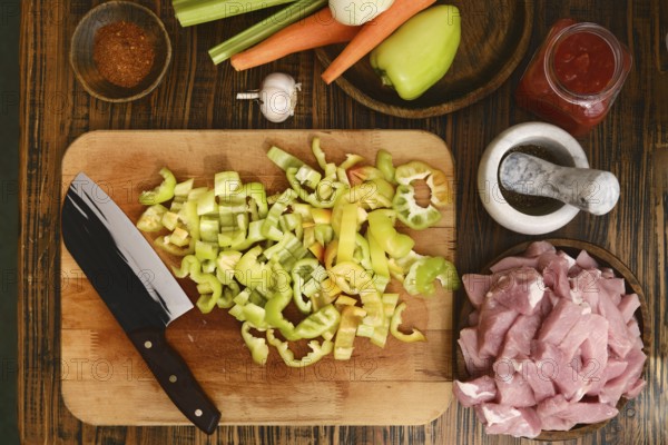 Freshly chopped green bell peppers, celery, and carrots are arranged on a wooden cutting board next to raw meat and spices, creating a vibrant setup for a home-cooked meal preparation