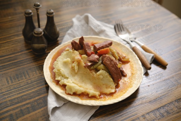 A plate filled with creamy mashed potatoes topped with tender pieces of meat and colorful vegetables is placed on a rustic wooden table