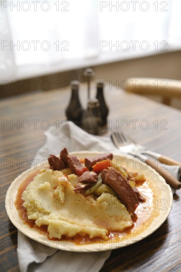 A hearty serving of creamy mashed potatoes topped with tender meat and vegetables on a rustic wooden table. Soft light illuminates the meal, enhancing the inviting atmosphere of the setting