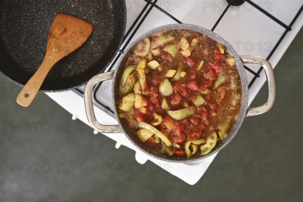 A pot filled with chopped vegetables and pork meat simmers on a stovetop. Fresh tomatoes, green peppers, and spices create a colourful and aromatic stew