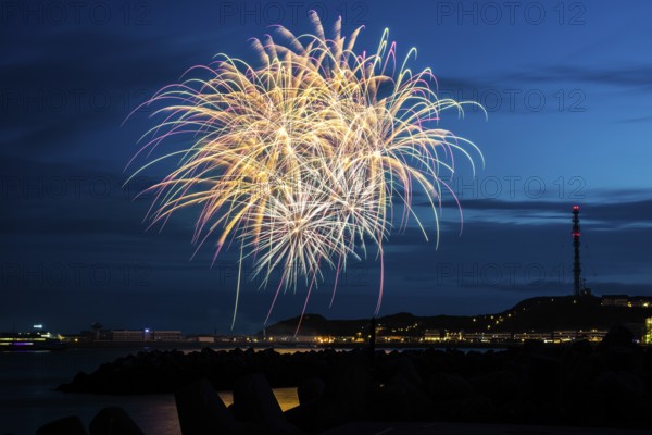 Atmospheric, splendid, intense fireworks with many illuminated sheaves for the Heligoland island festival, nocturnal lowlands with inland harbour, jetty and lobster shacks, on the right the uplands with transmission tower, transmitter, in front the groynes of the dune, reflecting water of the North Sea, blue night sky at the island festival in summer, symbolic image also for New Year's Eve, New Year's Eve fireworks, bright, colourful, many colourful traces of light, lively glitter, night, Heligoland Island, Schleswig-Holstein, Germany