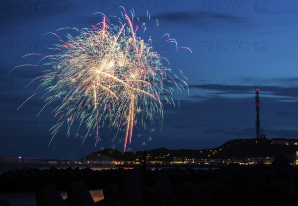 Brilliant, atmospheric fireworks display for the Heligoland island festival in red, blue and gold, nocturnal lowlands with inland harbour, jetty and lobster shacks, on the right the uplands with transmission tower, transmitter, in front the groynes of the dune, reflecting water of the North Sea, blue night sky at the island festival in summer, symbolic image also for New Year's Eve, New Year's Eve fireworks, bright, colourful, many colourful traces of light, sparks, lively glitter, night, Heligoland Island, Schleswig-Holstein, Germany