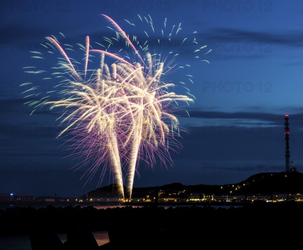 Spectacularly brilliant, atmospheric fireworks display for the Heligoland island festival in red, green and gold, the lowlands at night with the inland harbour, jetty and lobster shacks, on the right the uplands with the transmission tower, transmitter, in front the groynes of the dune, reflecting water of the North Sea, night sky at the island festival in summer, symbolic image also for New Year's Eve, New Year's Eve fireworks, bright, colourful, many colourful traces of light, sparks, lively glitter, night, island of Heligoland, Schleswig-Holstein, Germany