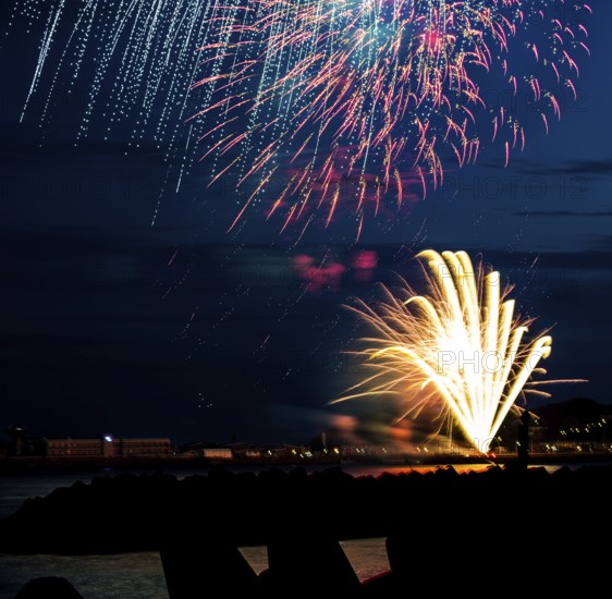 Brightly shining atmospheric fireworks for the Helgoland island festival in red, blue and gold over the nocturnal lowlands, inland port, jetty and lobster shacks, in front the stages of the dune made of tetrapods and the reflecting water of the North Sea, night sky at the island festival in summer, symbolic image also for New Year's Eve, New Year's Eve fireworks, bright, colourful, many colourful traces of light, sparks, lively glitter, night, island of Helgoland, Schleswig-Holstein, Germany