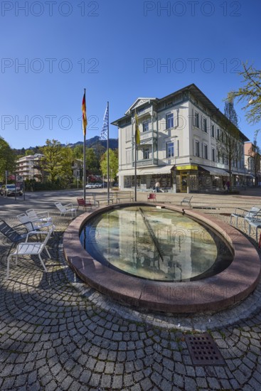 Wisbacherbrunnen, fountain, public chairs, flagpoles, flags, German flag, Bavarian flag, pedestrian zone, retail shops, buildings, trees, blue sky, cloudless, Wisbacherstrasse, Ludwigstrasse, Salzburger Strasse, Bad Reichenhall, Berchtesgadener Land district, Bavaria, Germany