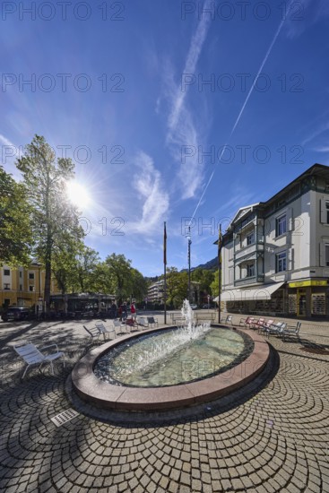 Wisbacherbrunnen, fountain, pedestrian zone, general architecture, cobblestones, public chairs, flagpoles, flags, trees, backlight of the sun, blue sky, cirrostratus clouds, intersection of Salzburger Straße with Wisbacher Straße and Ludwigstraße, Bad Reichenhall, district Berchtesgadener Land, Bavaria, Germany