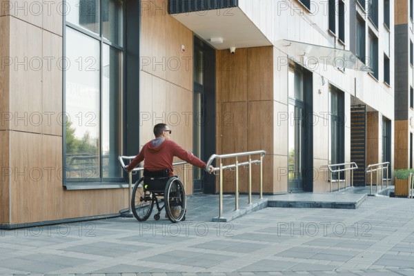 A person in a red hoodie navigates a wheelchair along a smooth ramp outside a contemporary structure. The sun is shining, highlighting the building's clean lines and wooden accents