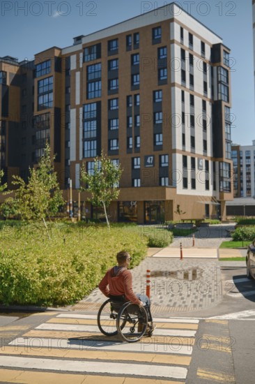 A man using a wheelchair navigates a crosswalk in a modern urban setting. The sun shines brightly, illuminating the well-designed buildings and landscaped greenery surrounding him