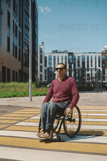 A man in a wheelchair navigates a crosswalk marked with yellow lines in a contemporary city environment. Modern buildings provide a backdrop under a bright blue sky