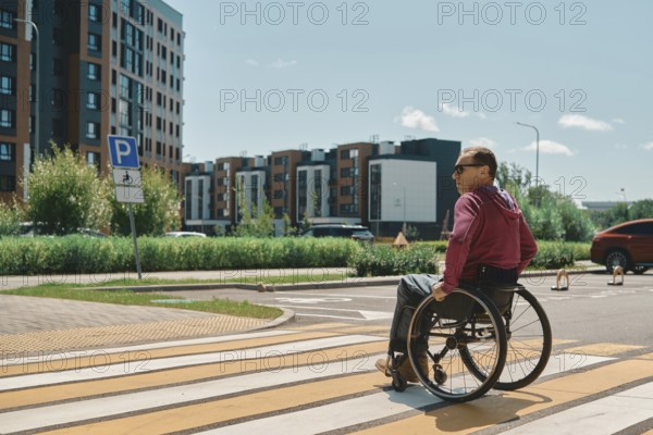 A man in a wheelchair is crossing a bright crosswalk on a sunny day. Surrounding him are modern buildings and greenery, showcasing a vibrant urban environment