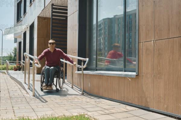 Individual in a wheelchair is utilizing a ramp to exit a modern building with large windows. The bright sun illuminates the exterior, creating a vibrant urban atmosphere