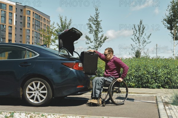 A person in a wheelchair is carefully placing a suitcase into the trunk of a parked car. The setting features modern buildings in the background and clear skies, suggesting a warm day