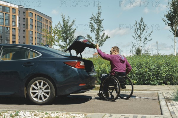 A man in a wheelchair is placing items into the trunk of a car in a bright urban area. The scene shows modern buildings and greenery, highlighting accessibility and independence on a clear day
