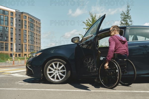 A man in a wheelchair is using his arms to propel himself into a parked vehicle with an open door. The backdrop features a contemporary apartment building under a clear blue sky
