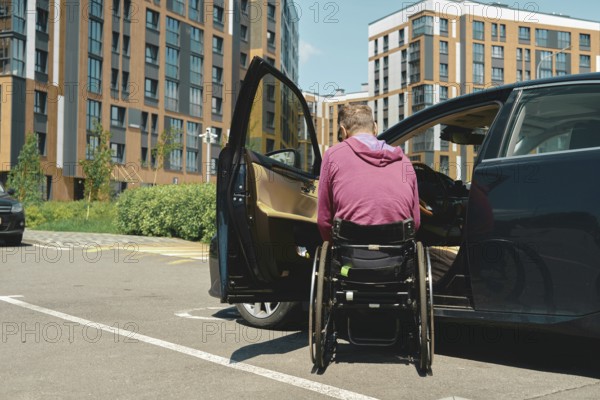 A man in a wheelchair is preparing to enter a car parked in a modern urban parking lot