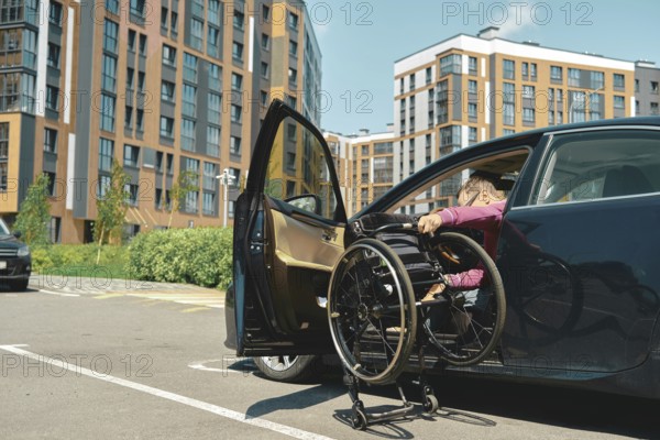 A person is maneuvering a wheelchair into a car parked in a contemporary urban area. A man unfolds a wheelchair to get into it from the car